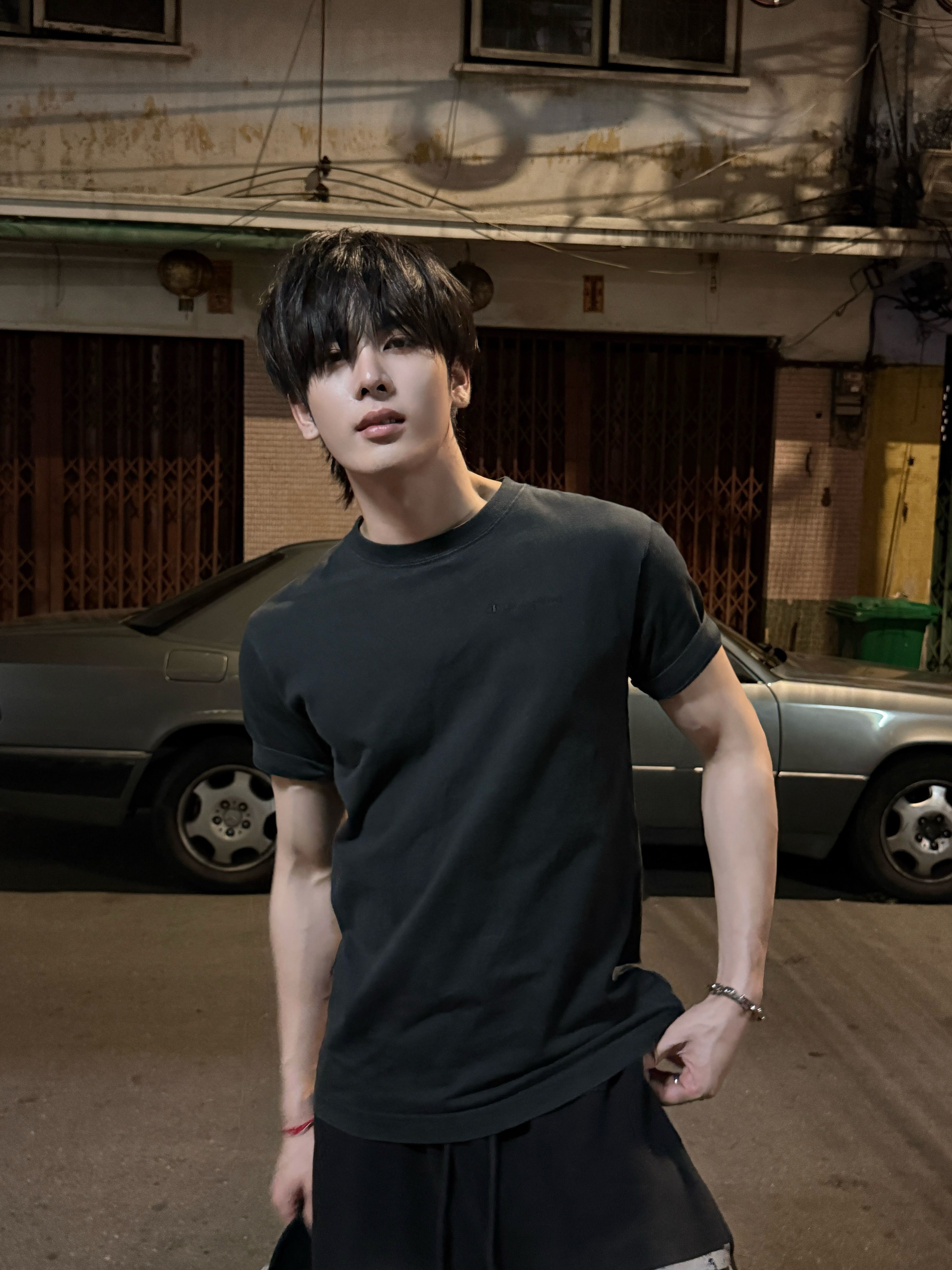 黒髪の青年が黒い半袖Tシャツとパンツを着て旧ビルの夜景でカメラを見つめている