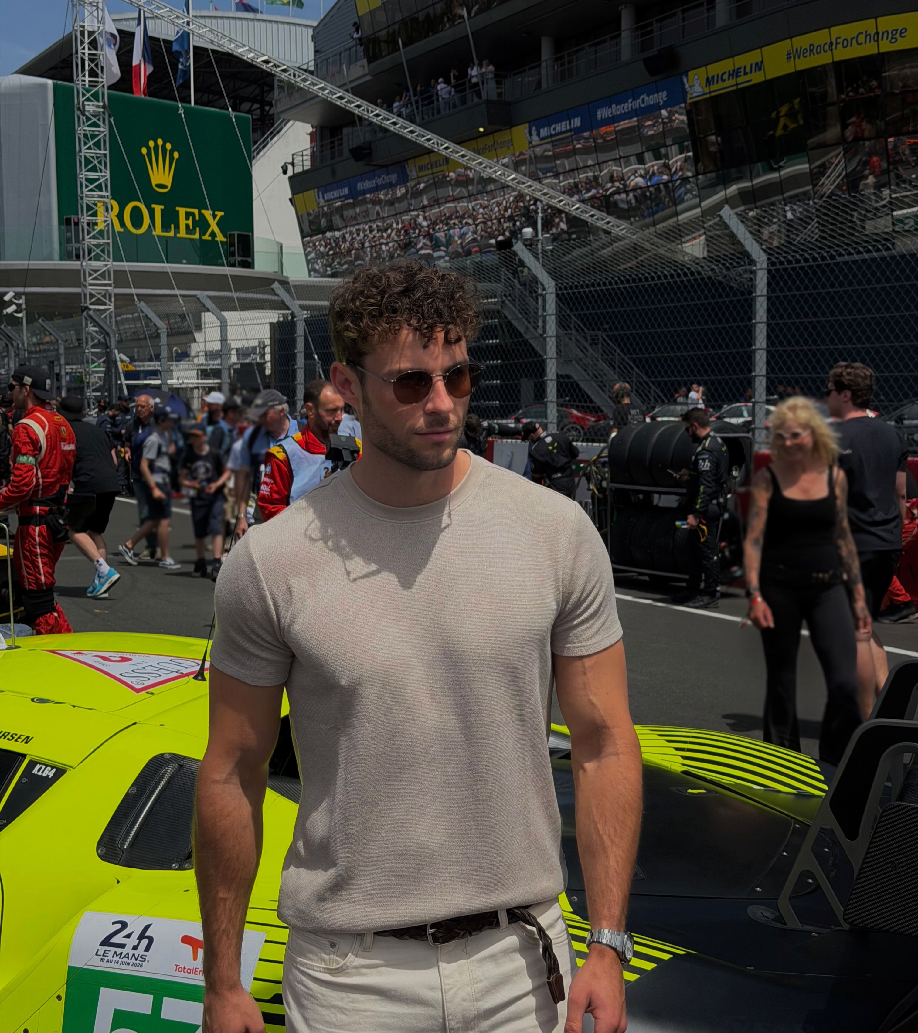 muscular young man wearing metal aviator sunglasses and light beige knit short sleeve t shirt with off white pants stands in le mans pit area