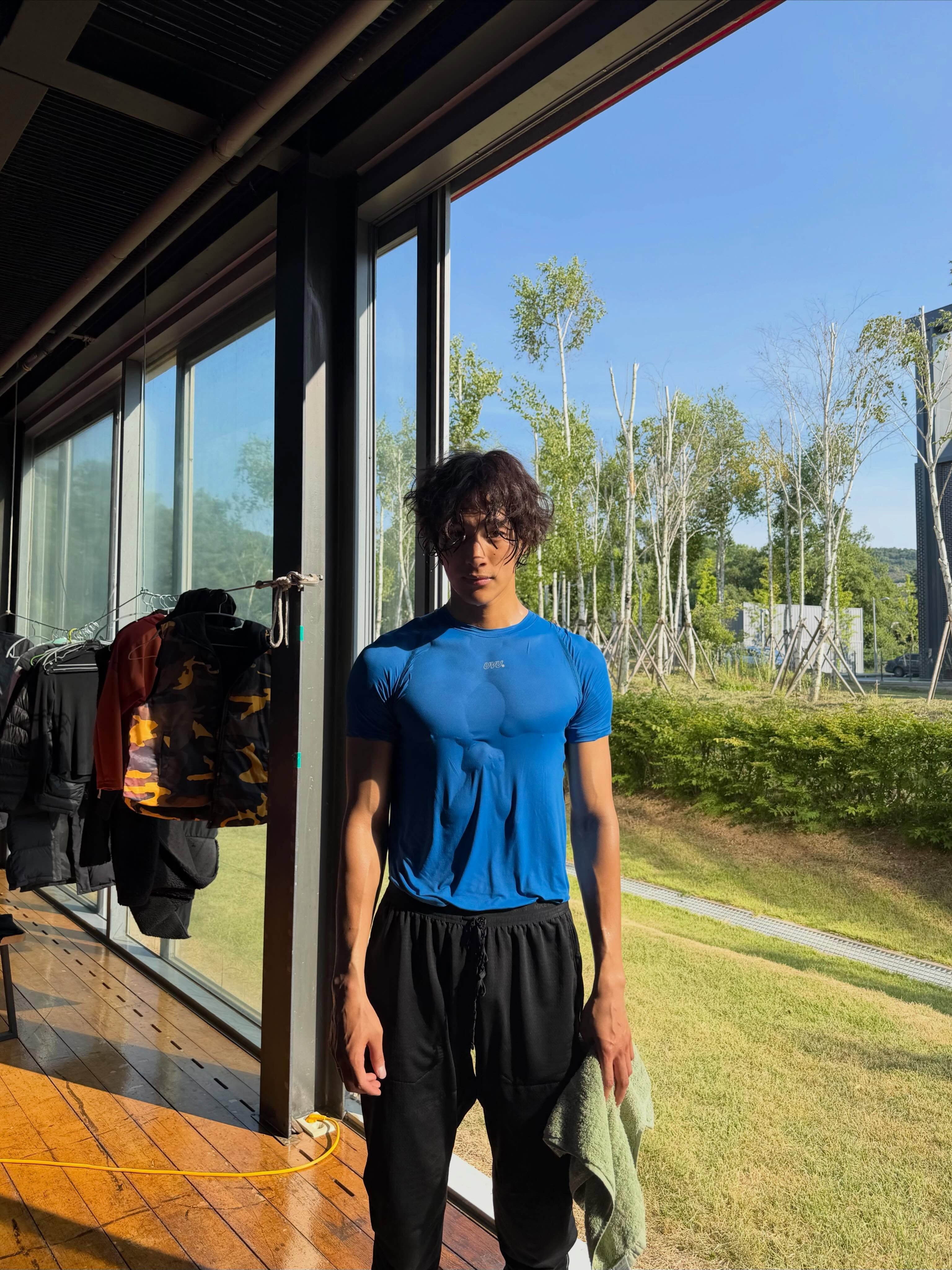 Jeune homme asiatique portant un t-shirt de sport bleu moulant et un pantalon noir ample debout près d'une fenêtre intérieure