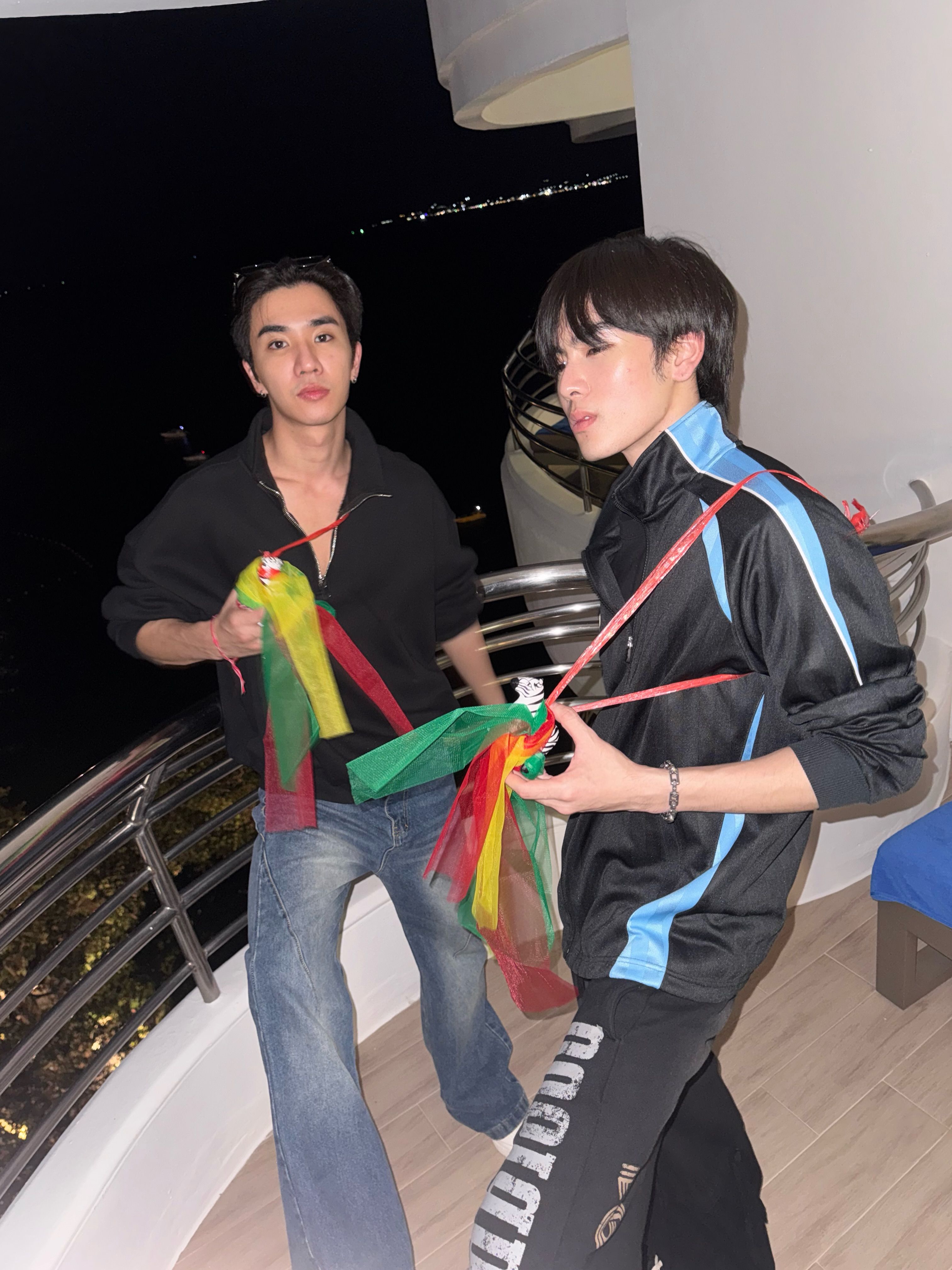 黒髪の青年が黒服の男性と夜のバルコニーでカラフルな人形を持って交流