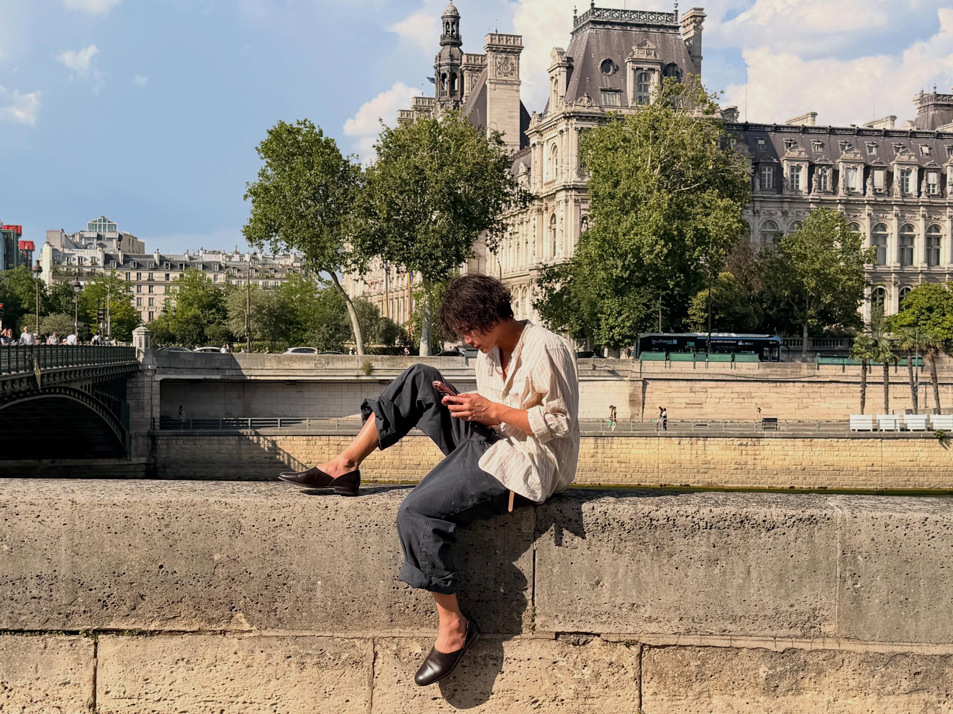 Asiatischer junger Mann trägt weißes gestreiftes Hemd und dunkle weite Hose sitzt am Flussufer in Paris und schaut auf sein Handy
