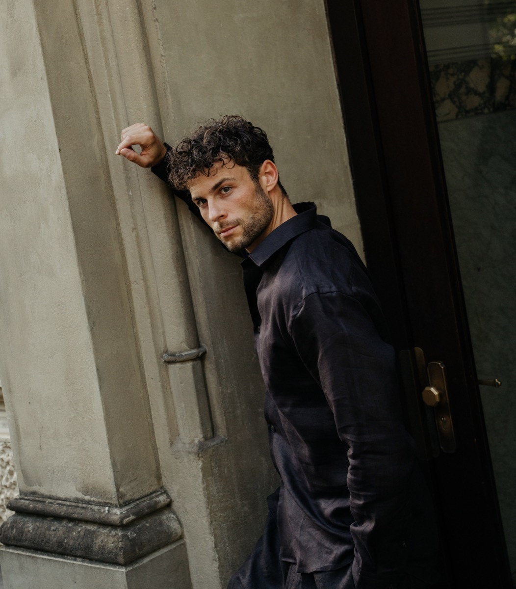 A young man in a dark navy silk shirt stands at the entrance of a stone building